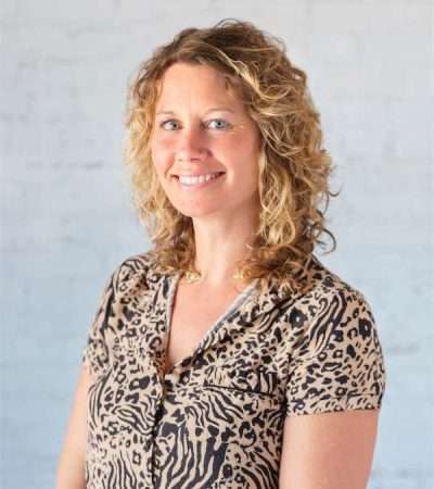 Stephanie Hertel, therapist in NYC, smiles at the camera. She has curly blonde hair and wears a short-sleeve animal print blouse, standing in front of a light-colored, textured background. Blue Canary Psychotherapy NYC