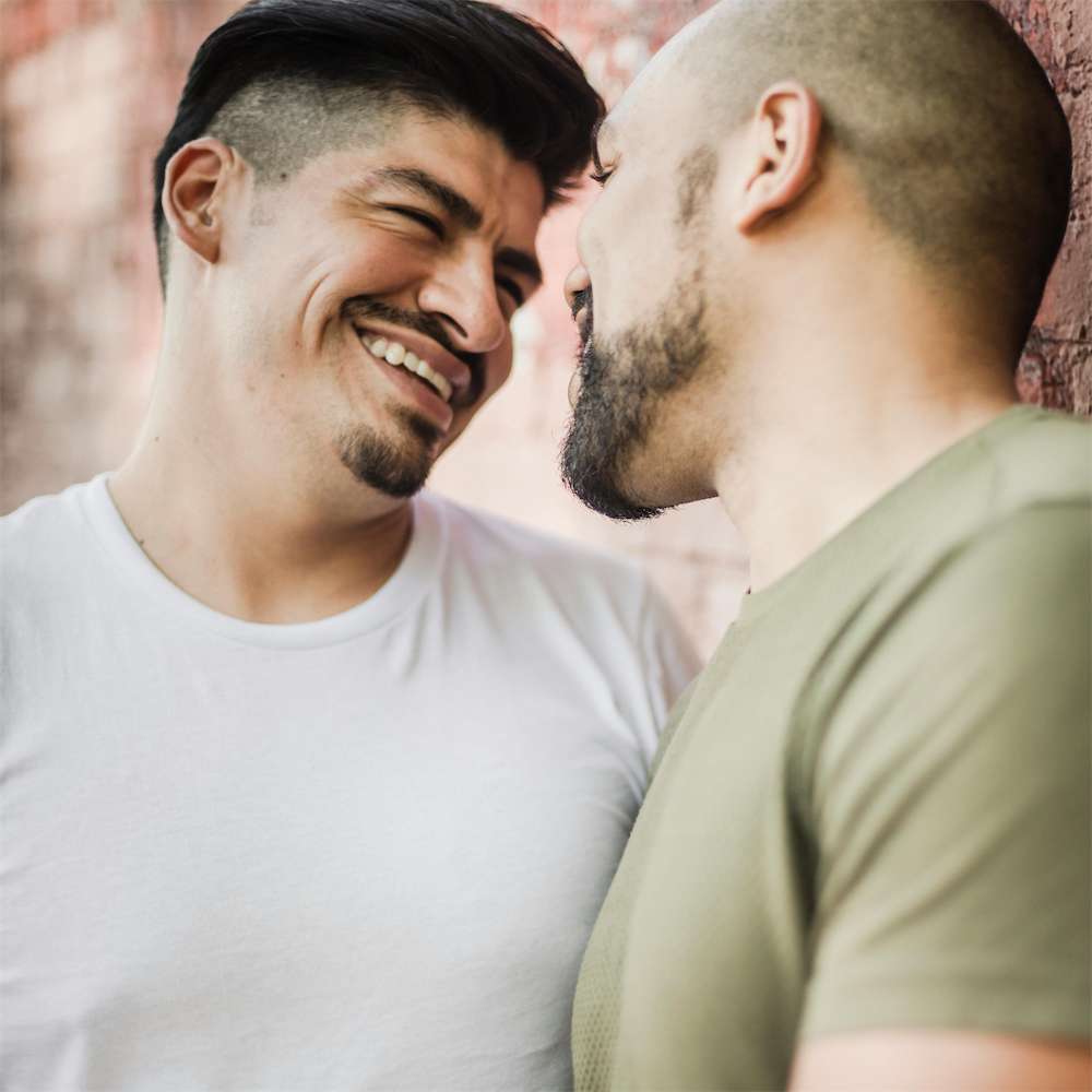 Two men stand close together, smiling and gazing into each others eyes. One wears a white shirt, the other a green shirt. Leaning against a brick wall, they share a moment of connection and joy, an embodiment of love that flourishes with the understanding nurtured in lgbtq+ couples therapy. Blue Canary Psychotherapy NYC