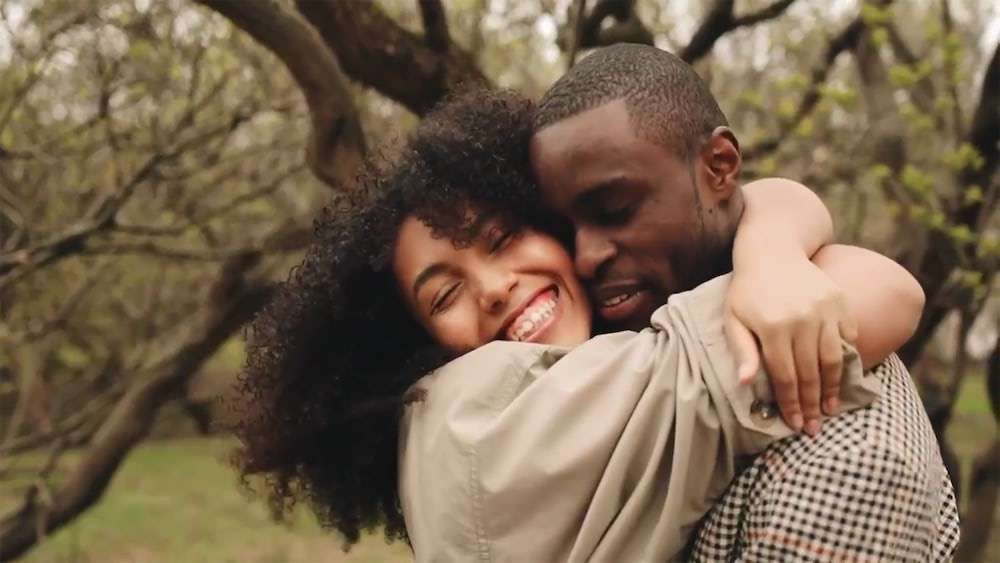 Una mujer sonriente con el pelo rizado abraza estrechamente a un hombre al aire libre entre árboles sin hojas. Ambos parecen felices y se abrazan cariñosamente, reflejando la conexión que a menudo se nutre a través de la terapia sexual que buscan las parejas de NYC. Blue Canary Psicoterapia NYC