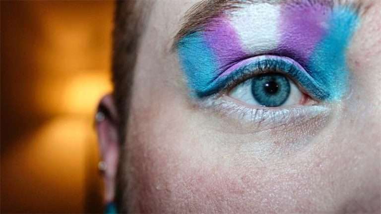 A close-up of a persons eye with bold, colorful makeup in blue, purple, and white shades, accentuating the eyelid and brow bone. The softly lit background reflects themes of self-expression and transgender mental health. Blue Canary Psychotherapy NYC