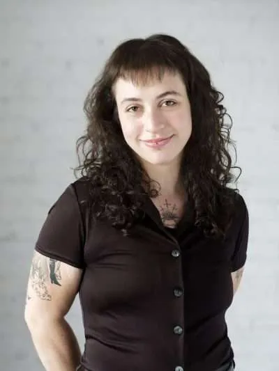 Talia Weinreb, therapist at Blue Canary Psychotherapy, smiling in a black button-up top in front of a white brick wall